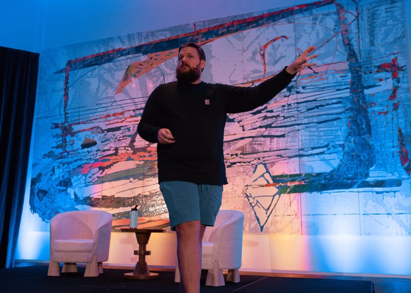 Speaker in a black sweatshirt and shorts pointing across the stage in front of a large abstract painting