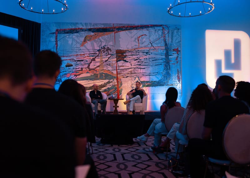 Guests watching two speakers in armchairs, with a large white logo block lit at the side
