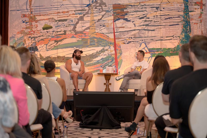 Two speakers in white armchairs talking before seated guests and a colorful painted wall