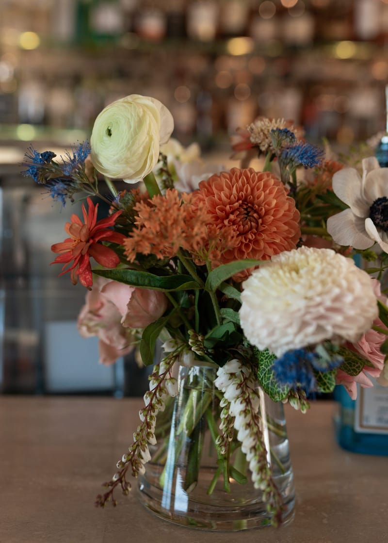 Dahlias, ranunculus and anemones in a glass vase on a bar lined with bottles