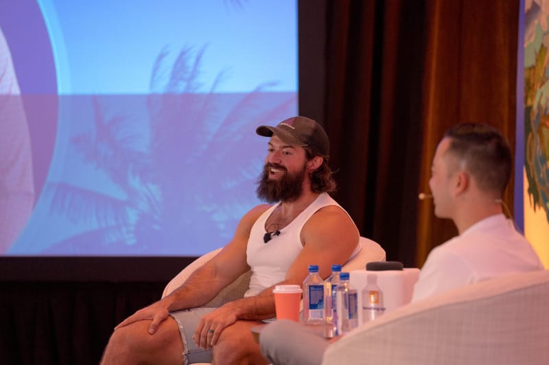 Two speakers in armchairs, one in a cap and tank top, with water bottles beside them