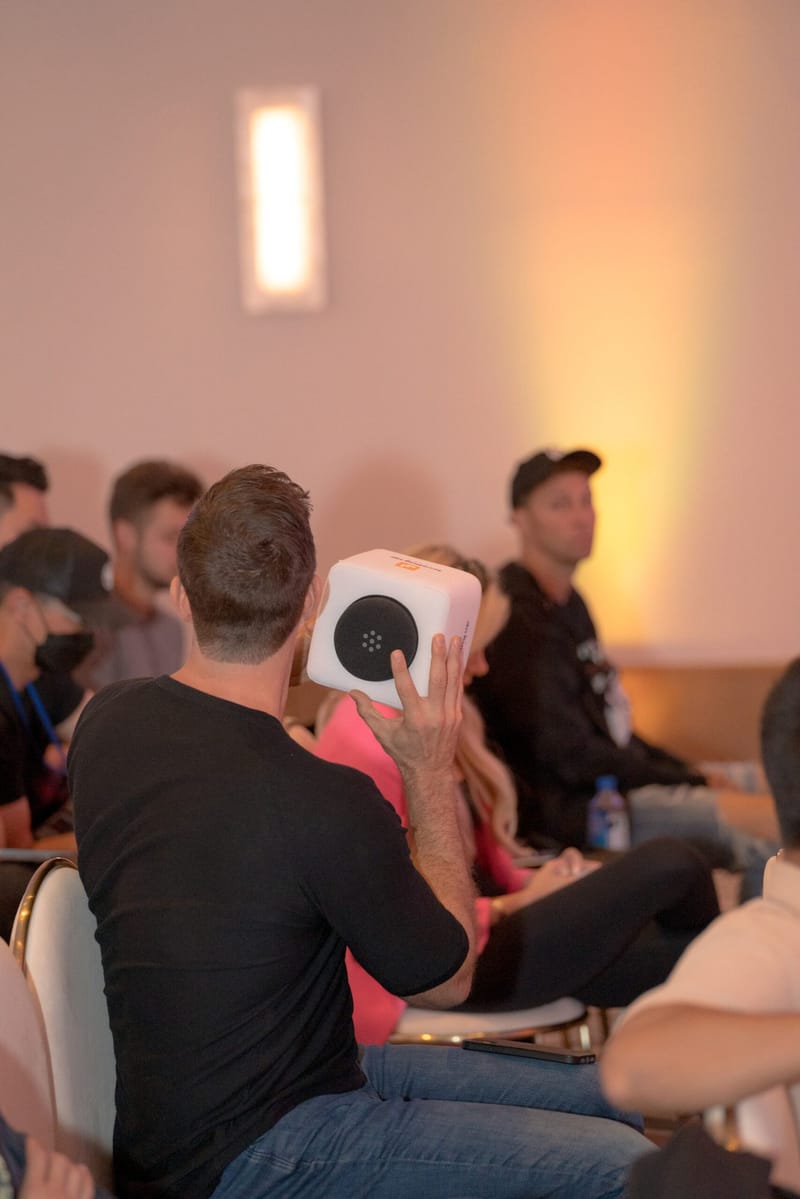Guest seen from behind raising a white cube microphone among seated guests