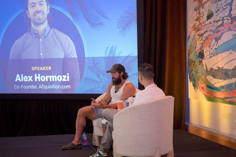 Two speakers in armchairs under a screen reading Speaker, Alex Hormozi, Co-Founder, Acquisition.com