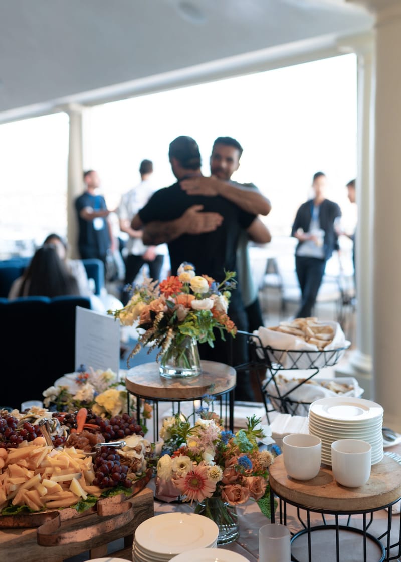 Grazing table with cheese, grapes, plates and mugs as two guests hug on a terrace behind