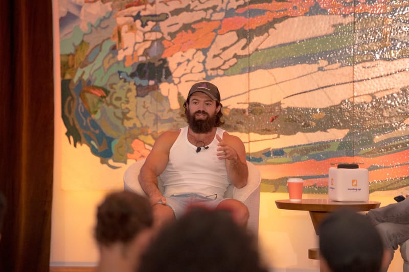 Speaker in a cap and white tank top gesturing from an armchair, a cube microphone beside him