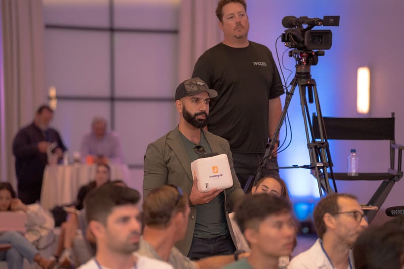 Guest standing with a white cube microphone beside a video camera on a tripod
