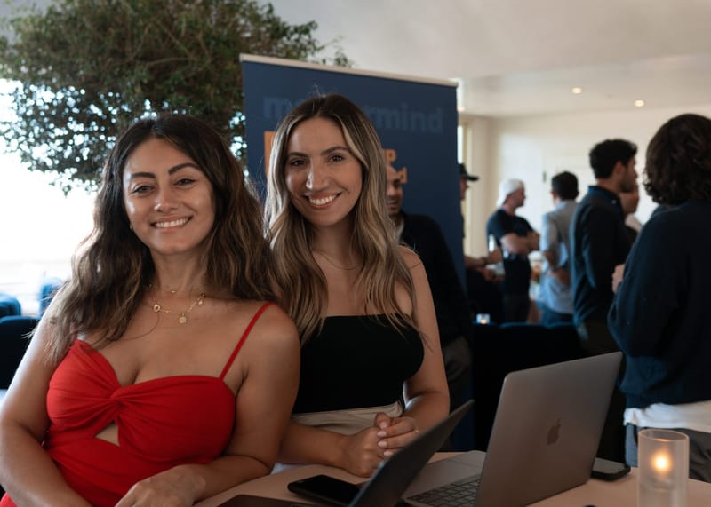 Two guests smiling at a table with laptops beneath an olive tree