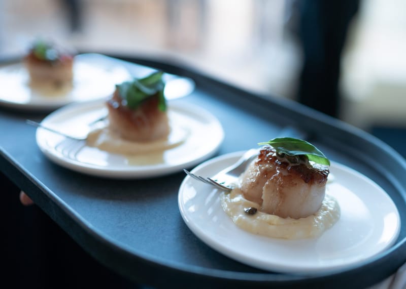 Seared scallops with basil on small white plates carried on a black tray
