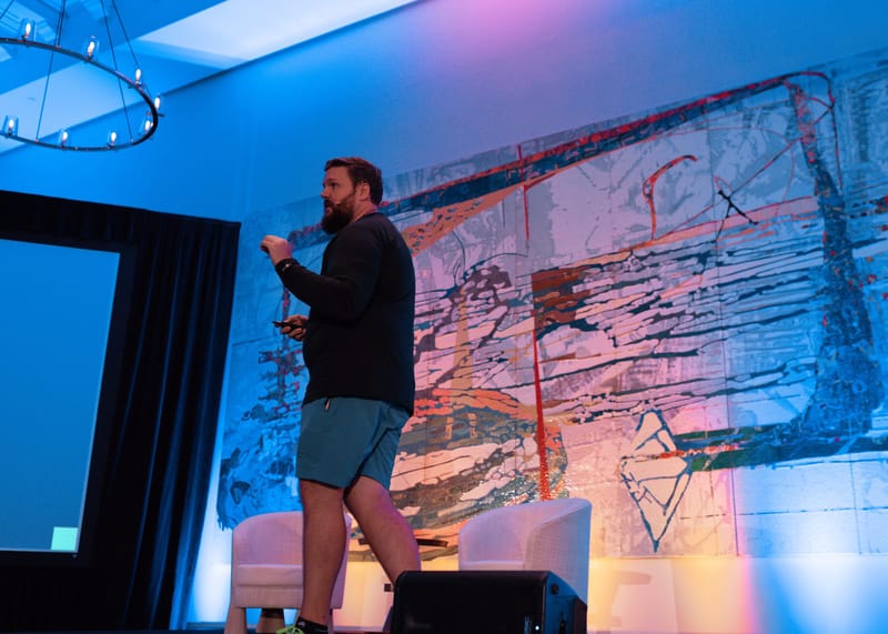 Speaker in shorts walking the stage in front of the painted wall and armchairs