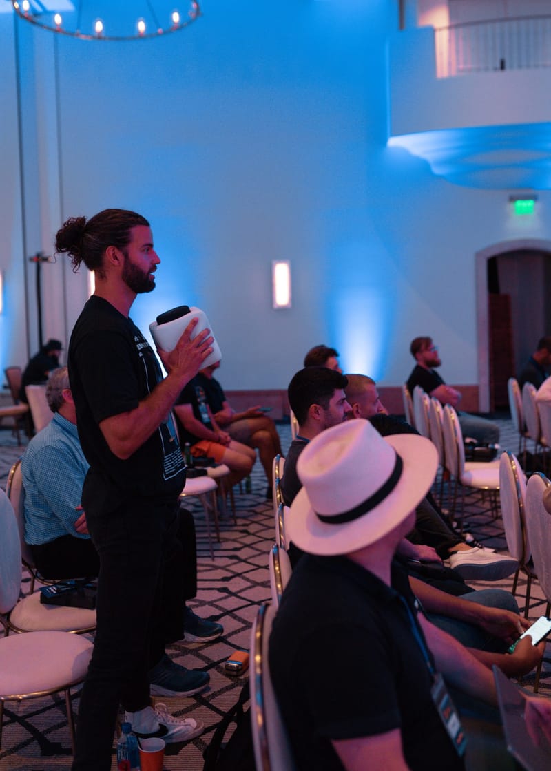 Guest standing to speak into a cube microphone beside a seated guest in a white hat