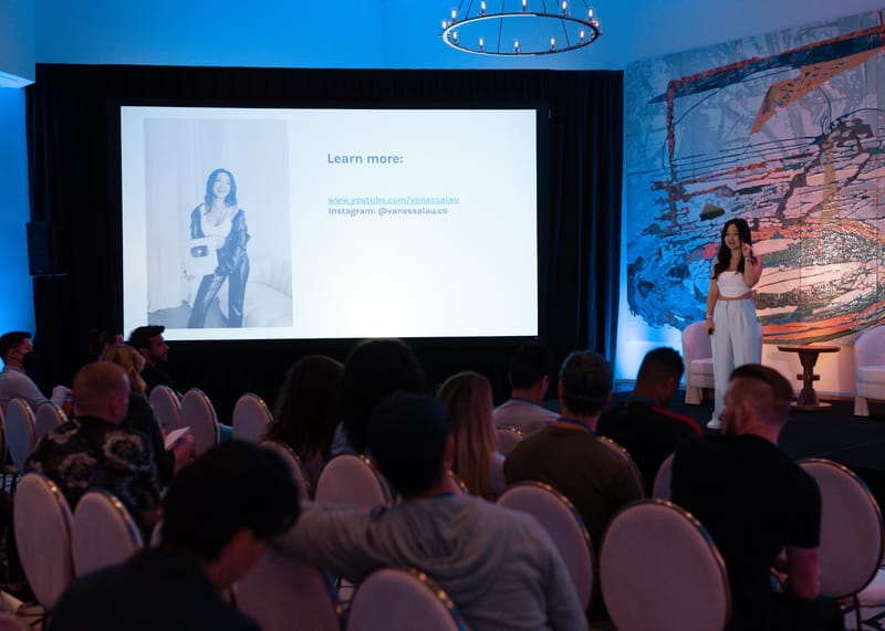 Speaker in white on stage beside a slide reading Learn more, facing a seated audience
