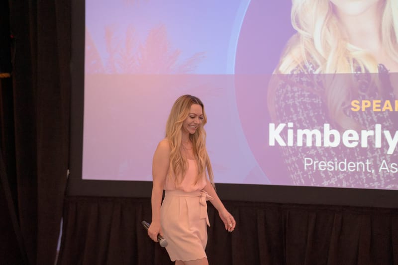 Speaker in a pink jumpsuit walking the stage under a screen reading Kimberly and President