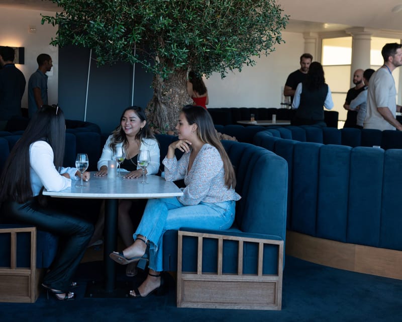 Three guests at a marble table in a navy velvet banquette under an olive tree