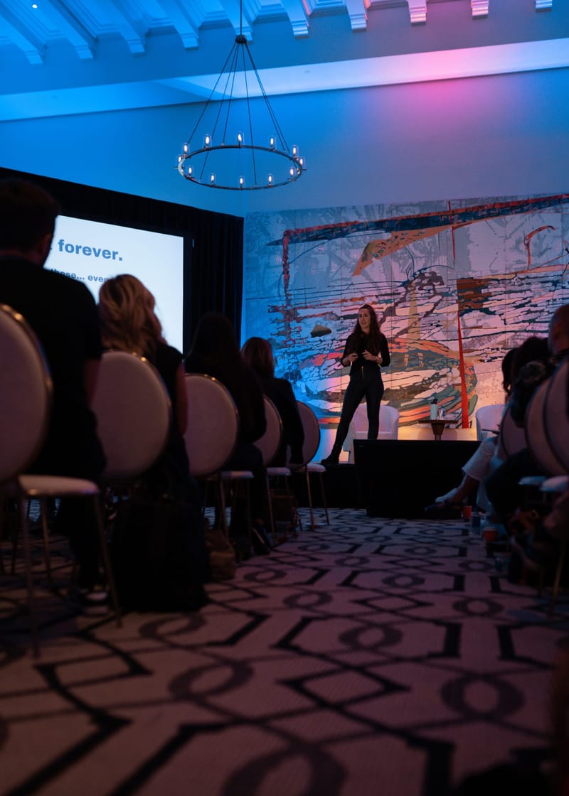 Aisle view to a speaker on stage, with a screen reading forever. at left