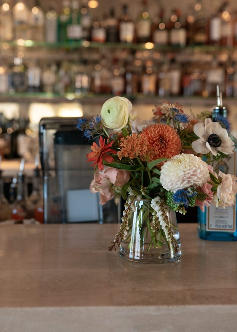 Orange dahlias and white ranunculus in a glass vase on a bar in front of shelves of bottles