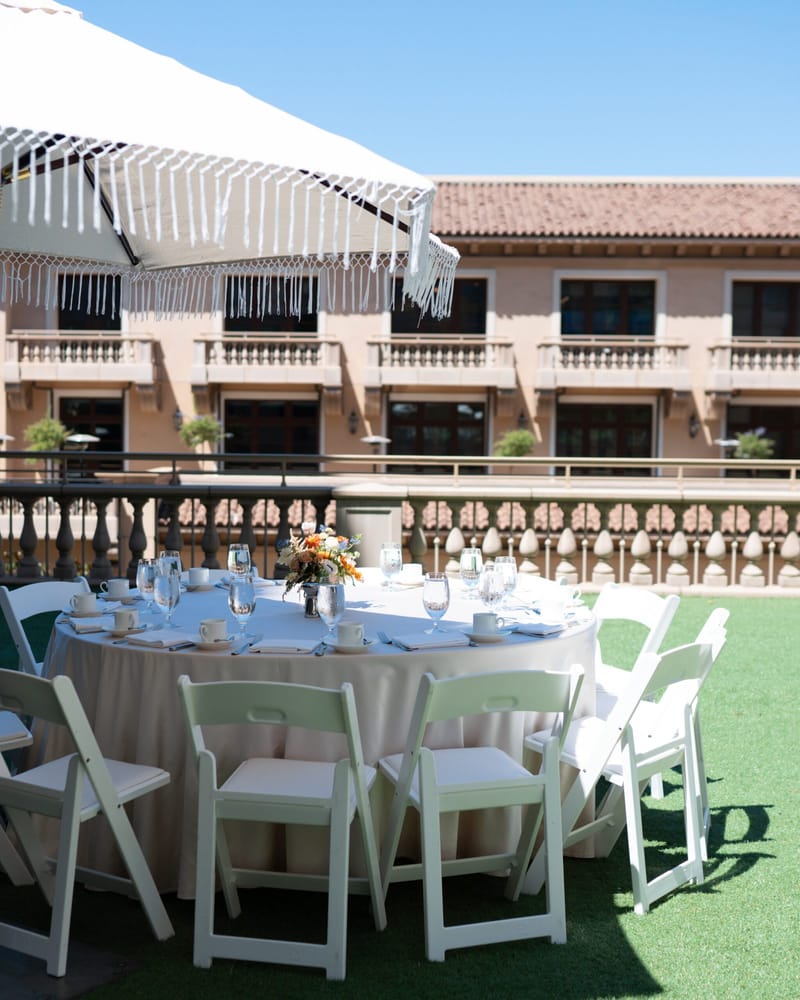 Round table set with white folding chairs on a lawn beside a fringed umbrella