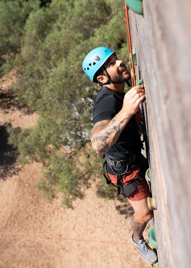 Guest in a blue helmet and harness climbing handholds on a wooden pole