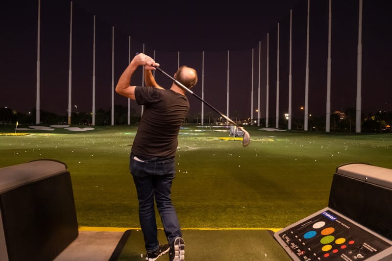 A guest swinging a golf club in a floodlit driving range bay at night