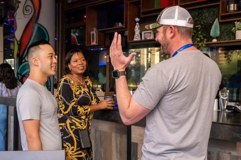 Three guests talking at a bar, one gesturing, beside a colorful painted mural