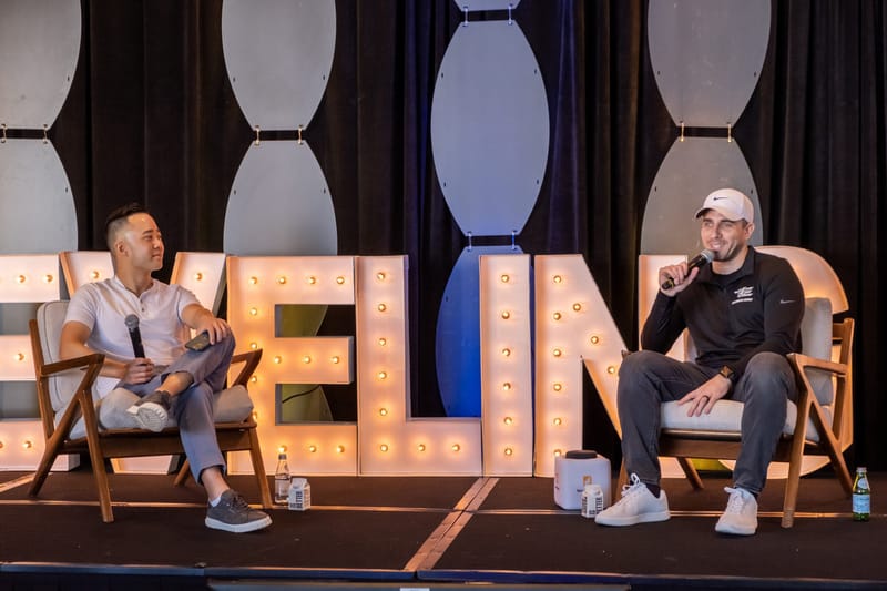 Two speakers seated in armchairs in front of LEVELING UP marquee letters, one holding a microphone
