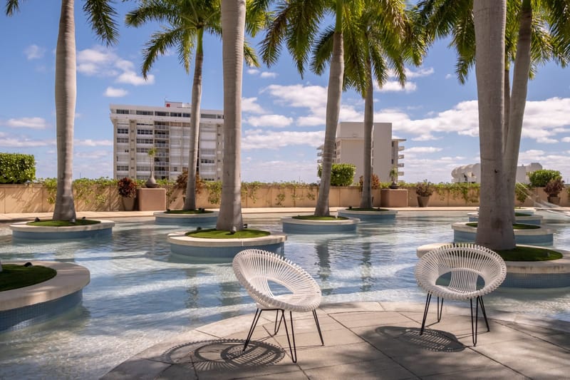 White chairs beside a shallow pool with palm trees in round planters under a blue sky