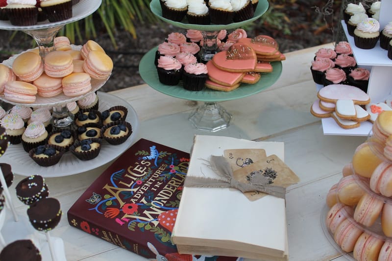 Dessert table of macarons, pink frosted cupcakes, heart cookies and an illustrated Alice's Adventures in Wonderland book