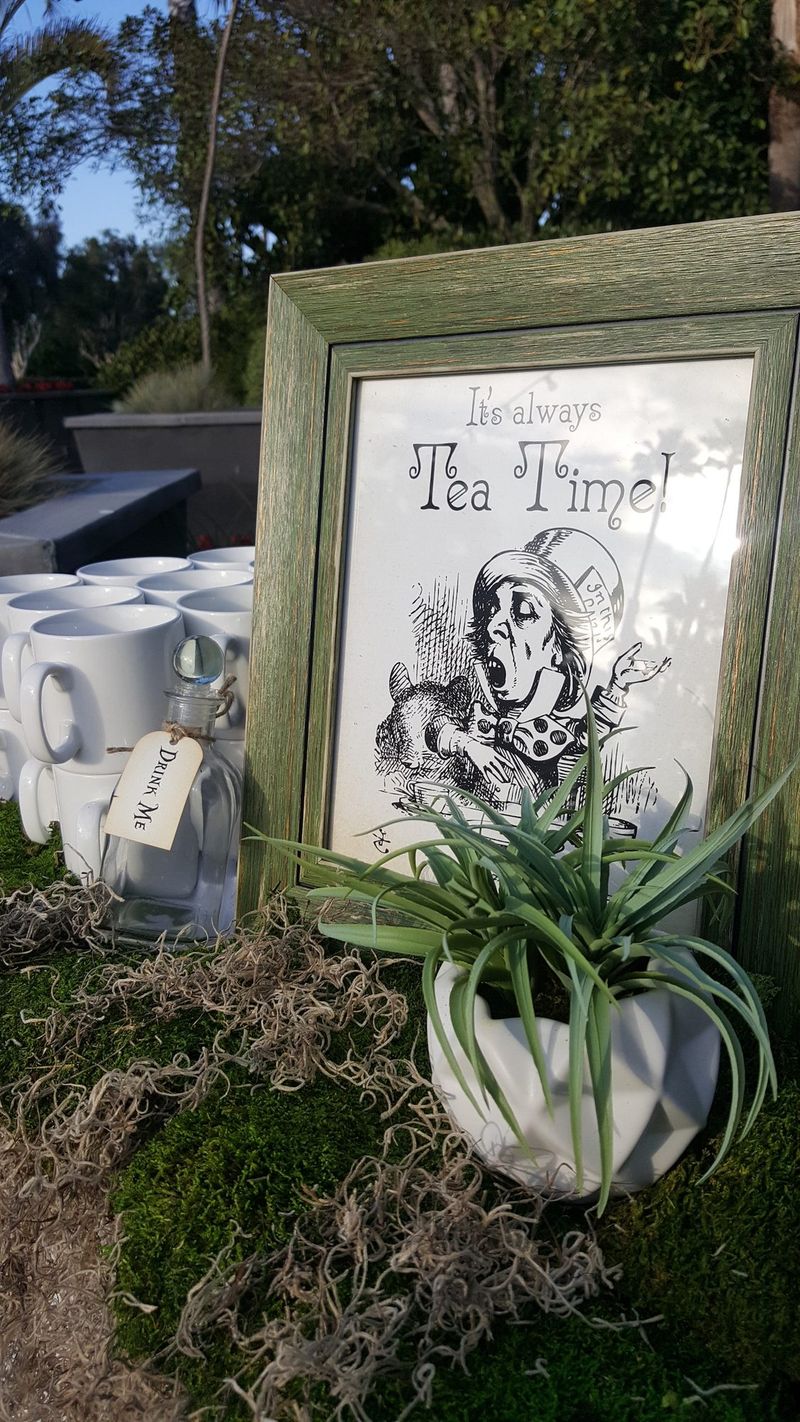 Framed Mad Hatter print reading It's always Tea Time! beside white mugs, a Drink Me bottle and an air plant