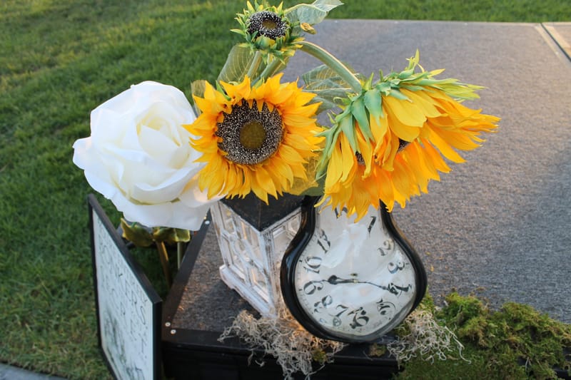 Sunflowers and a white rose beside a drooping clock on a moss-covered stand