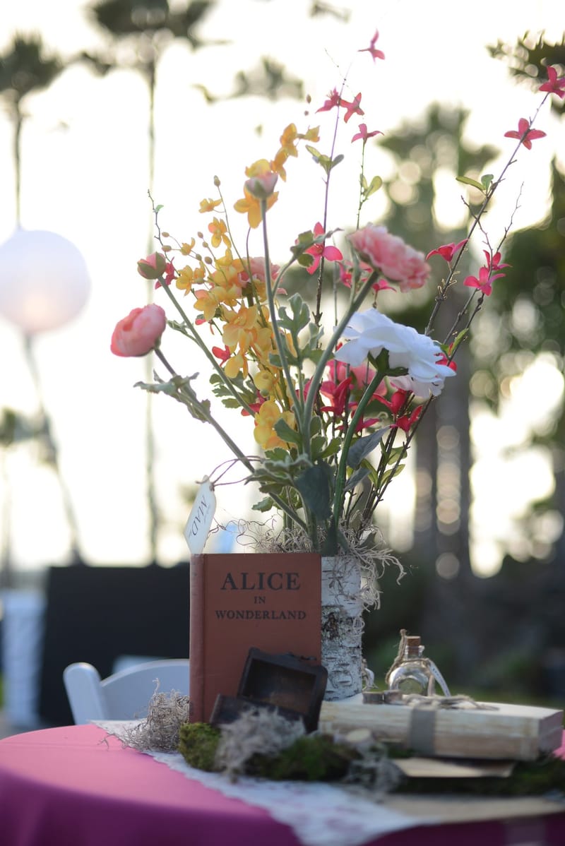 Wildflowers in a vase beside a red Alice in Wonderland book on a pink table, palm trees behind
