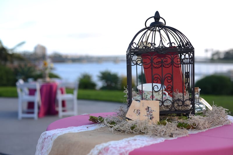 Black birdcage with a teacup and a small tag on a pink and burlap table, the water behind