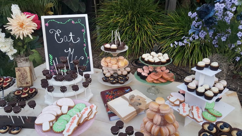 Dessert spread with an Eat Me chalkboard, a macaron tower, cupcakes, cake pops and cookies among flowers