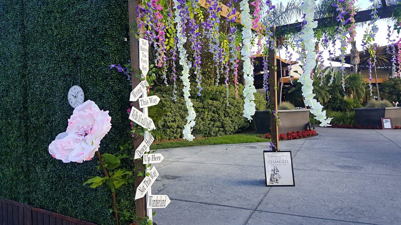 Wooden pergola hung with purple and white florals beside arrow signs, a giant pink flower and a wall clock
