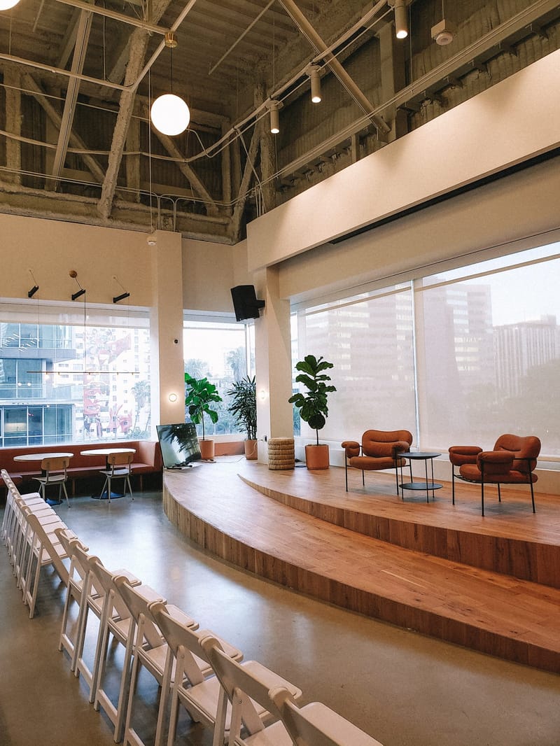 Empty hall with a curved wooden stage, orange lounge chairs, rows of white folding chairs and tall windows