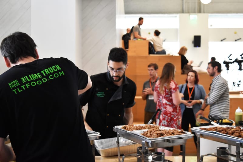 Two caterers, one in a The Lime Truck shirt, serving hot food from chafing dishes while guests talk behind them