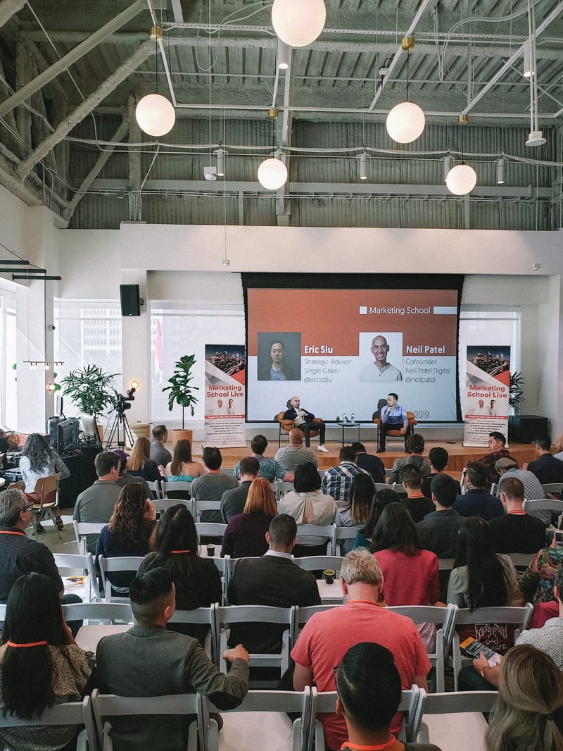 Full audience in white folding chairs facing the stage, the Eric Siu slide and Marketing School Live banners