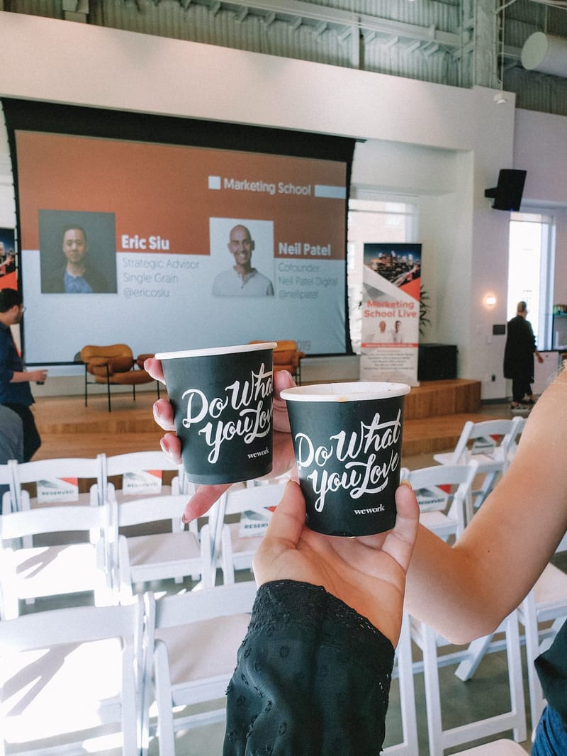 Two hands holding black coffee cups printed Do What You Love and wework above rows of reserved chairs