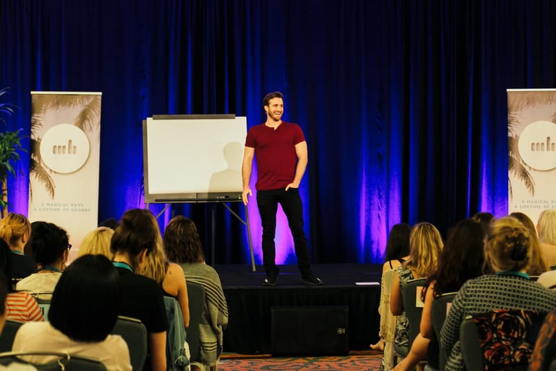 A speaker in a maroon T-shirt beside a flip chart on a stage with blue curtains and palm print banners
