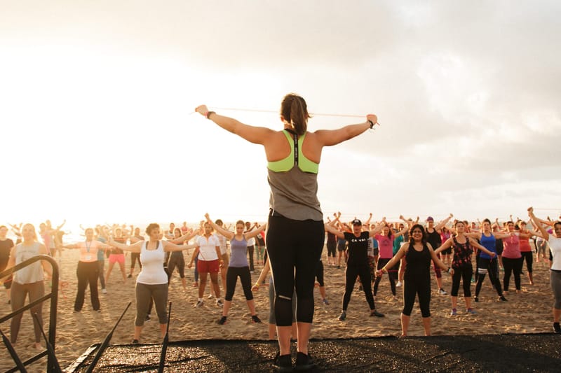 A woman on raised steps holding a band overhead as a crowd exercises on the beach at sunrise