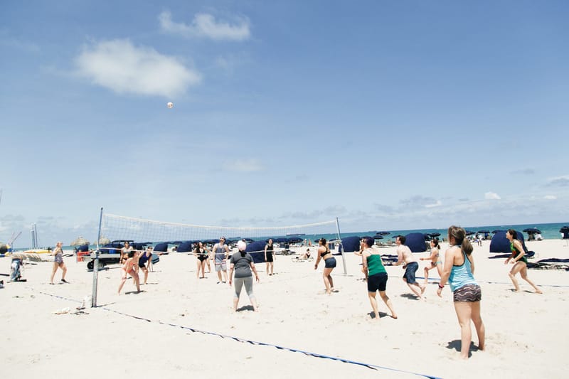 Guests playing volleyball on a wide sandy beach with blue cabanas behind