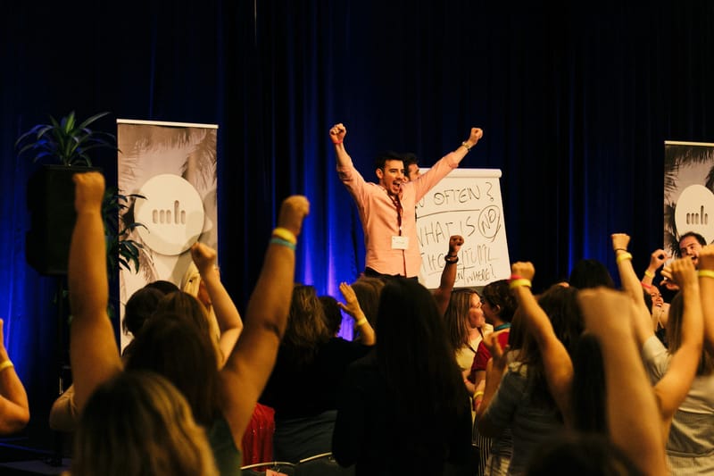 A man with a name badge cheering with both arms raised beside a flip chart as the crowd raises fists