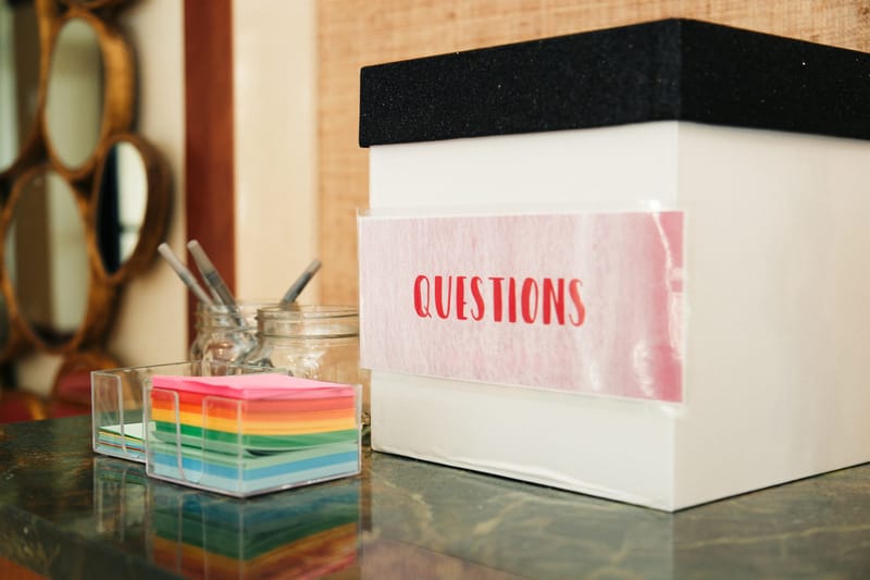 Box labeled Questions beside a stack of rainbow sticky notes and a jar of pens