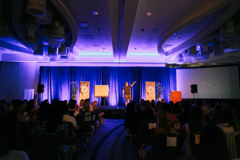 Wide view of a purple-lit room with a speaker on stage between flip charts and banners