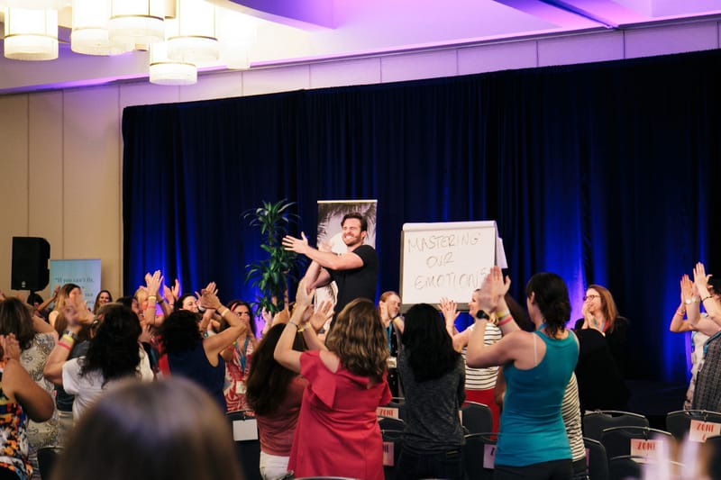 A speaker laughing beside a flip chart reading Mastering our emotions as guests clap overhead