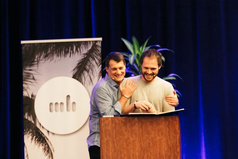 Two men laughing together at a wooden podium beside a palm print banner with the retreat logo