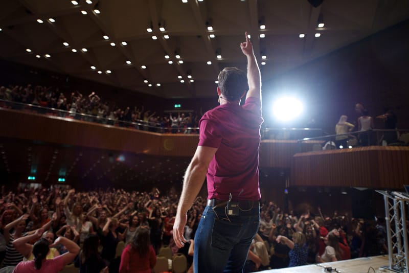 A speaker at the edge of the stage pointing upward in a spotlight, facing a full hall and balcony