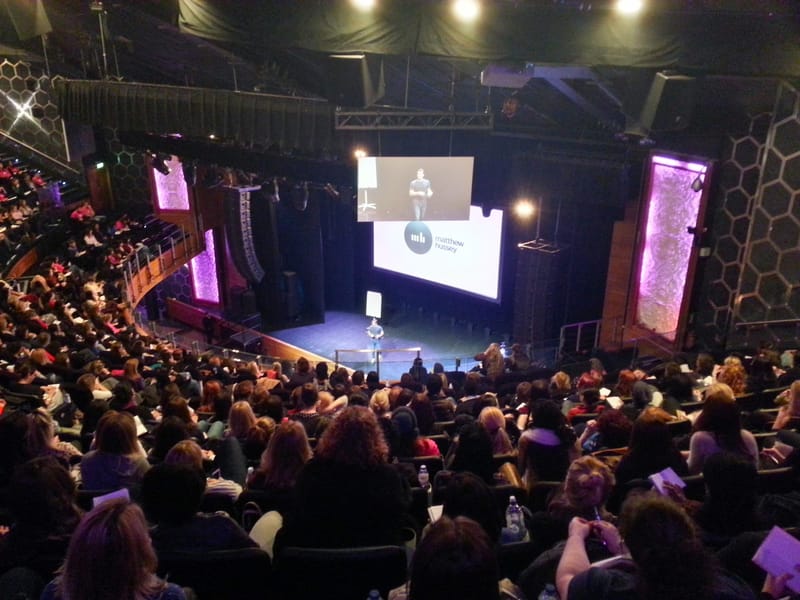 Theater seen from the balcony, a crowd facing a screen reading matthew hussey beside purple-lit wall panels