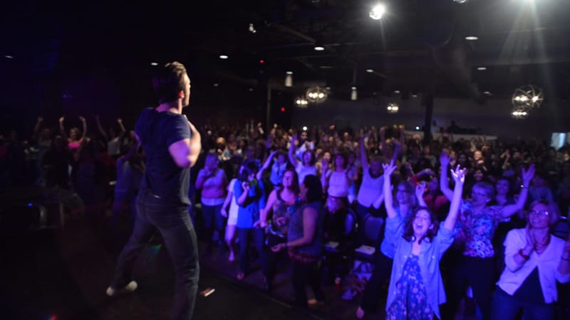A speaker on stage facing a crowd with raised arms under purple lighting