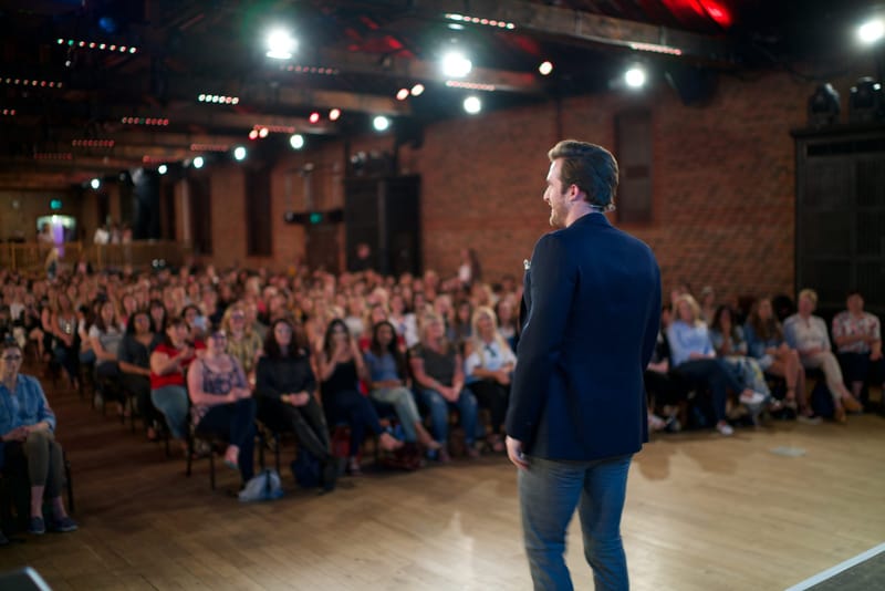 A speaker in a navy blazer on a wooden stage looking out at a seated crowd in a brick hall