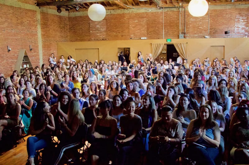 Smiling audience seated close together below white paper lanterns in a brick-walled hall