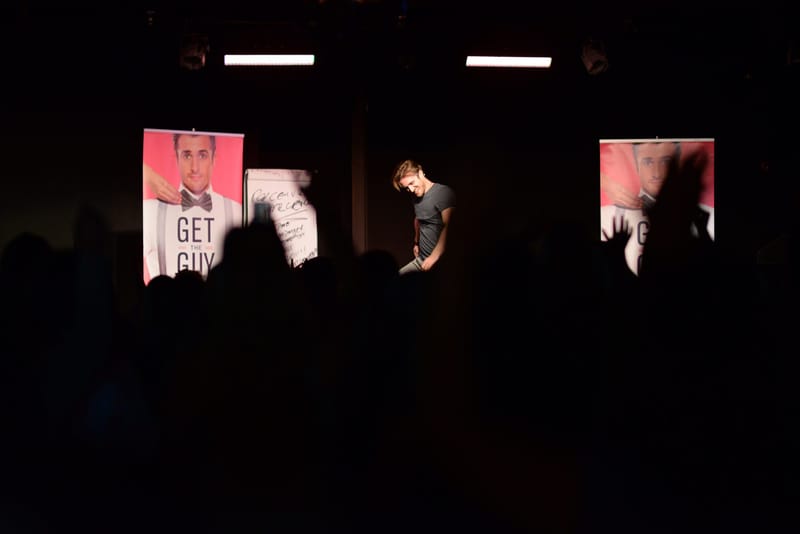 A speaker on a dark stage between Get The Guy posters and a flip chart, raised hands in silhouette